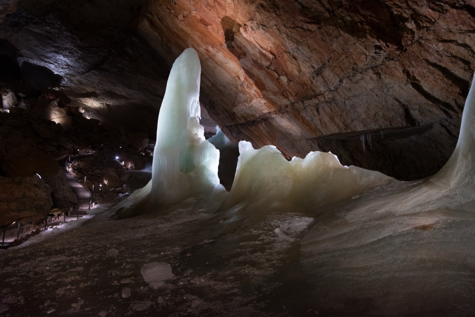 Lit columns and walls of ice inside a cave, with a stairway and path in the background